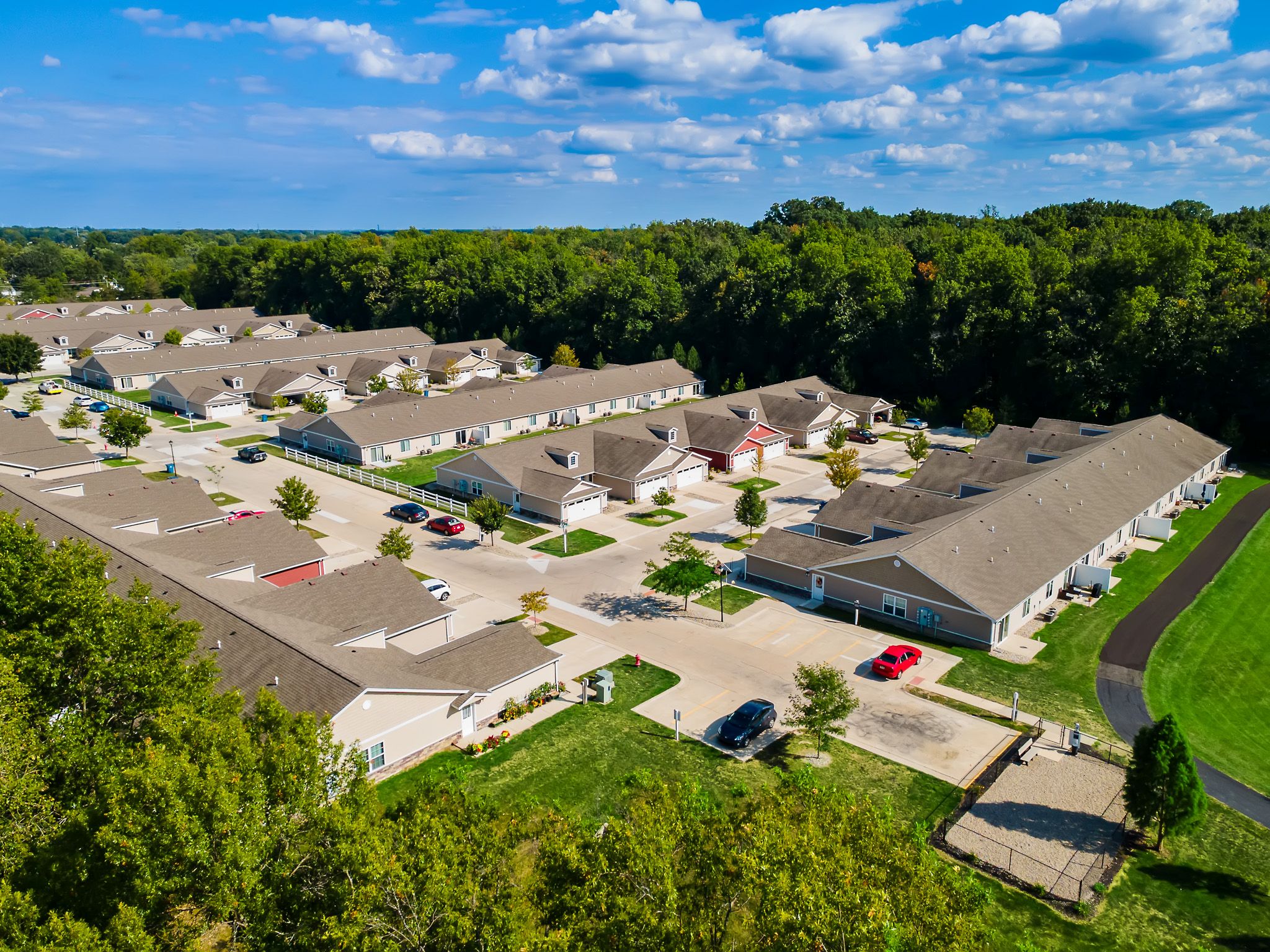 Aerial view of a residential neighborhood with single-story homes, driveways, parked cars, green lawns, and trees in the background under a partly cloudy sky.