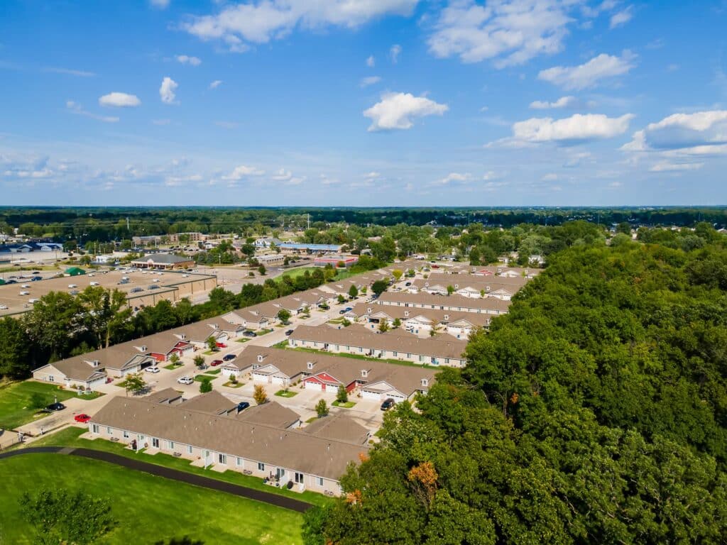 Aerial view of a residential neighborhood with rows of houses next to a wooded area under a partly cloudy sky.