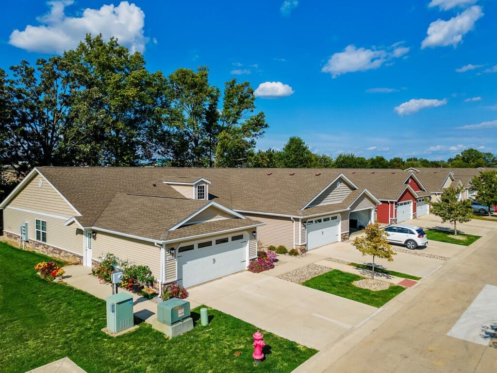 Row of attached suburban houses with driveways, garages, manicured lawns, and cars parked outside on a sunny day with blue sky and scattered clouds.