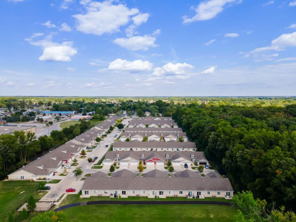 Aerial view of a suburban residential complex bordered by a forested area on one side and commercial buildings on the other, under a partly cloudy sky.
