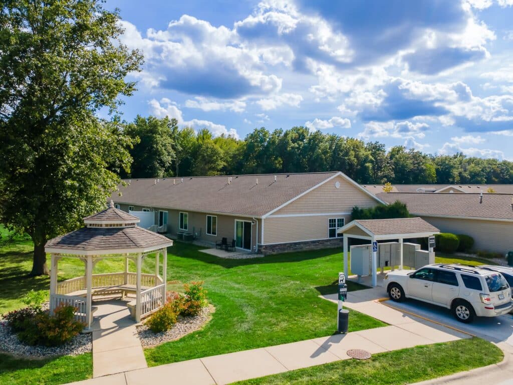 Single-story residential building with a gazebo on a landscaped lawn, accessible parking spaces, and a white SUV parked nearby under a partly cloudy sky.