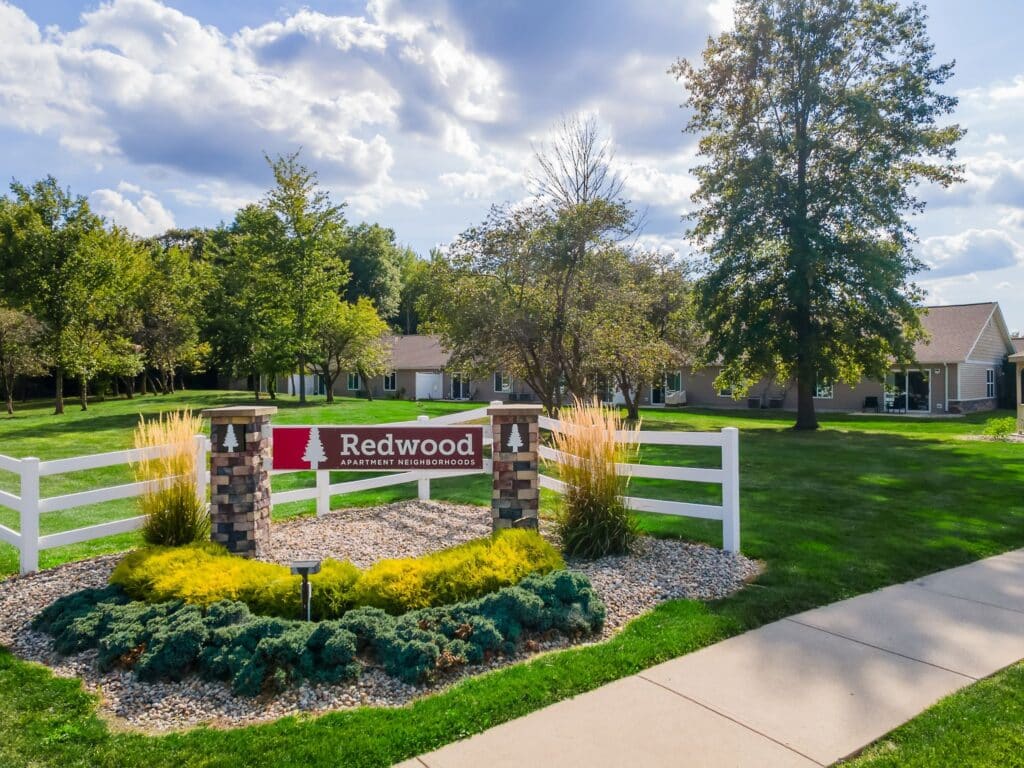 A landscaped entrance sign for Redwood Apartment Neighborhoods stands on a lawn with trees and ranch-style buildings in the background under a partly cloudy sky.