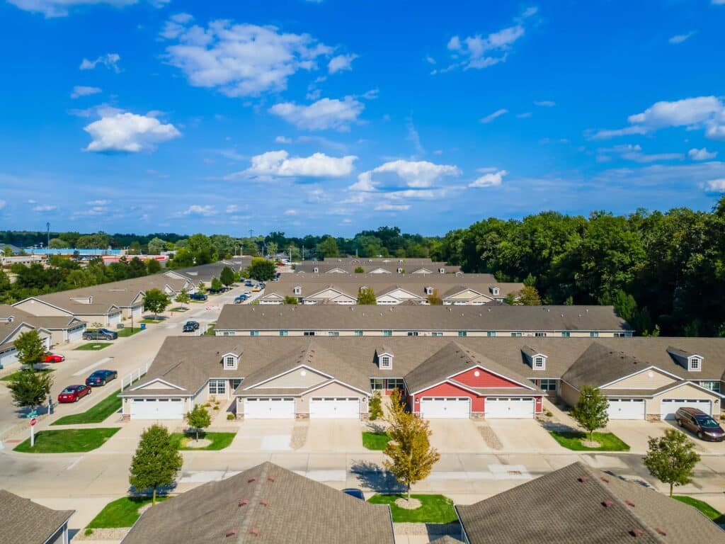 Aerial view of a suburban neighborhood with rows of single-story houses, cars parked in driveways, and trees bordering the area under a blue sky with scattered clouds.