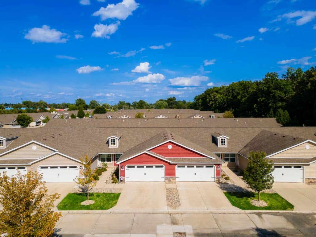 A row of attached suburban houses with beige and red exteriors, each with a garage and driveway, trees, and a clear blue sky above.