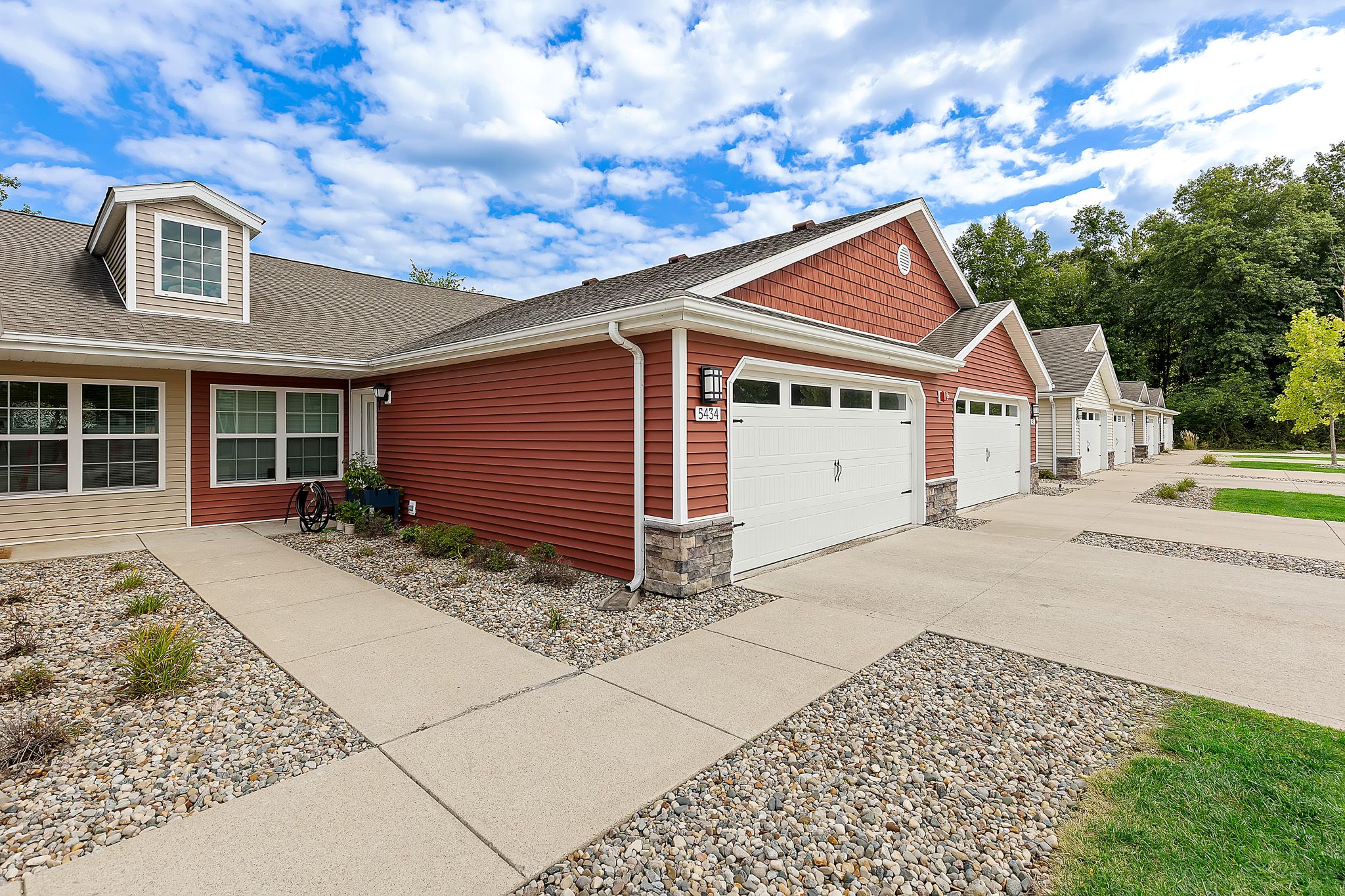Single-story red and beige townhouses with double garages, stone accents, and landscaped rock gardens along a wide concrete driveway under a partly cloudy sky.