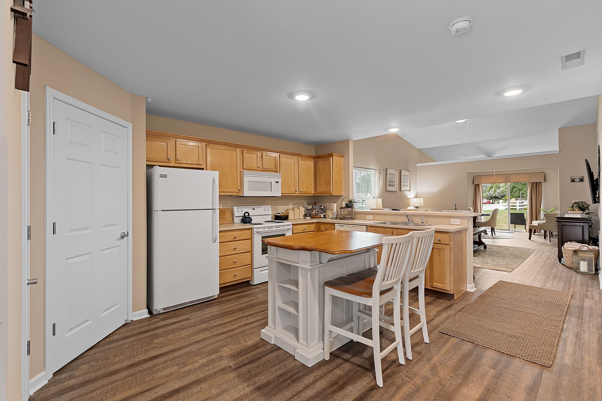 Open kitchen with wood cabinets, white appliances, a center island with two chairs, and view of a connected living area with sliding glass doors to the outside.