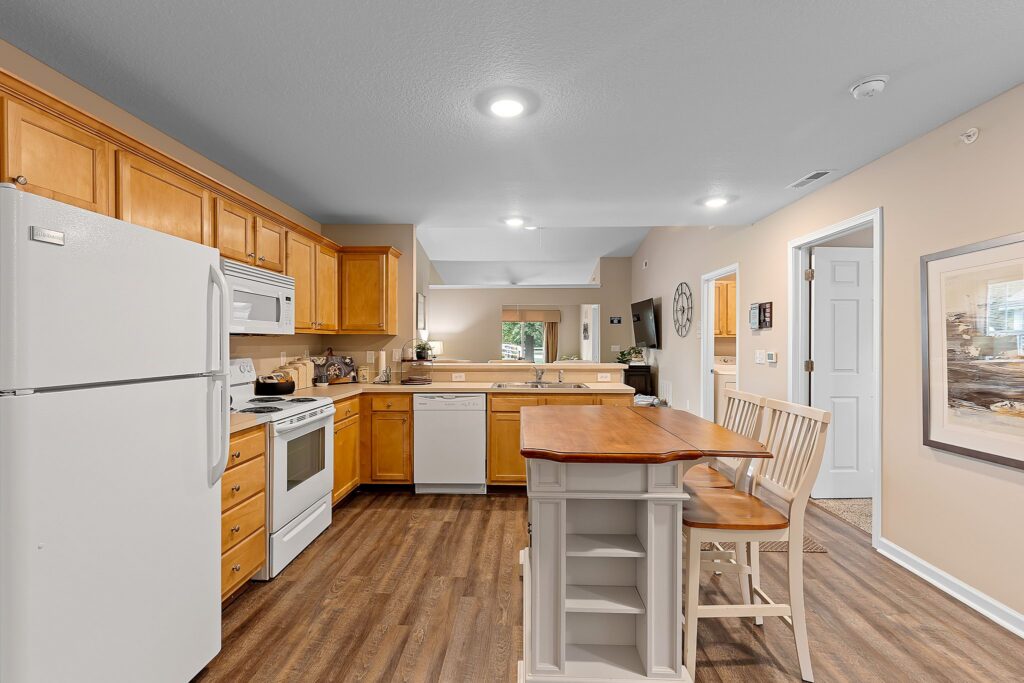 A kitchen with light wood cabinets, white appliances, an island with two stools, wood flooring, and neutral-colored walls.
