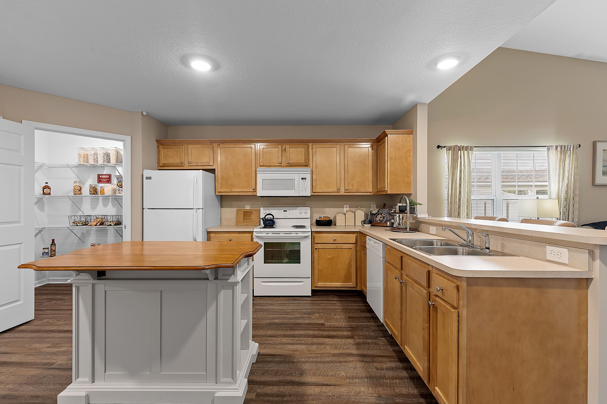 Modern kitchen with light wood cabinets, white appliances, a central island, double sink, and open pantry shelves; window with curtains lets in natural light.