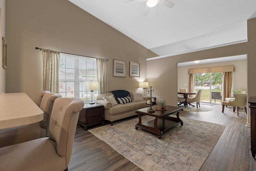 A spacious living room with a beige sofa, coffee table, area rug, and two windows; a dining area with a round table and chairs is visible in the background.