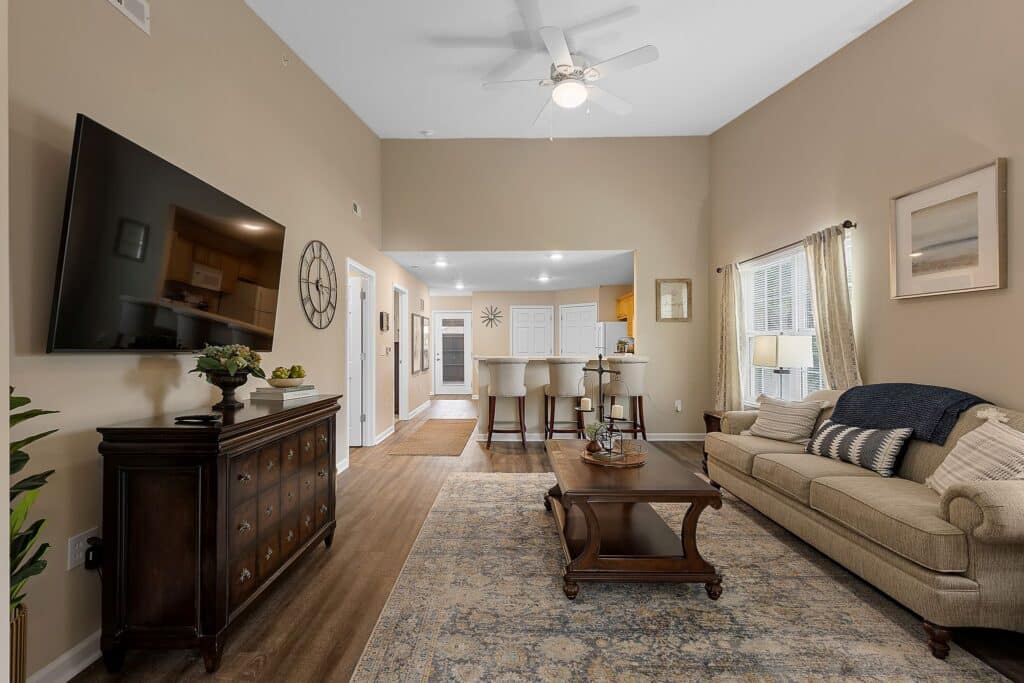 Spacious living room with beige walls, a mounted TV, wooden furniture, area rug, sofa with pillows, and a kitchen with bar stools in the background. Ceiling fan and large window visible.