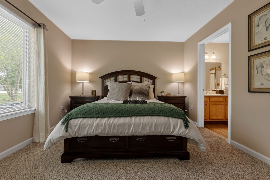A neatly made bed with a green blanket and pillows sits between two lamps in a carpeted bedroom; a window is on the left and an open door reveals a bathroom on the right.