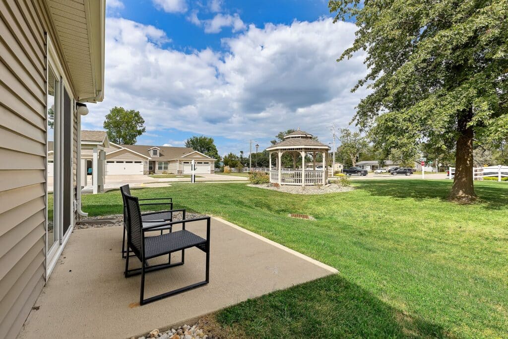 Two black patio chairs on a concrete porch overlook a grassy yard with a white gazebo, trees, and nearby houses under a partly cloudy sky.