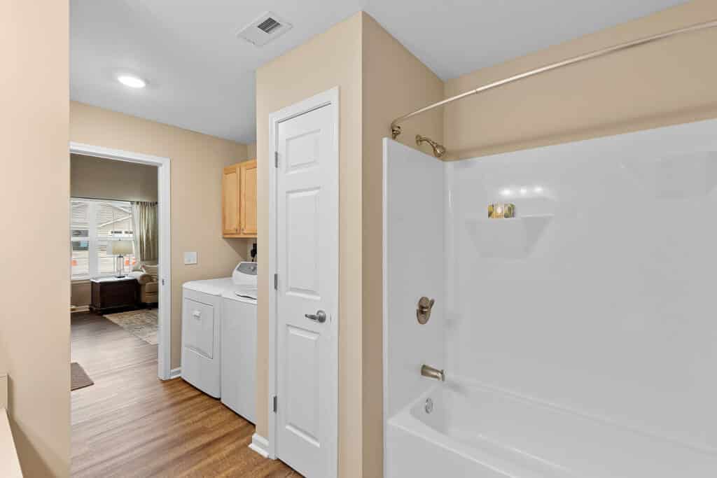 A bathroom with a white bathtub and shower, adjacent to a laundry area with a washer and dryer, and a door leading to another room with a window.