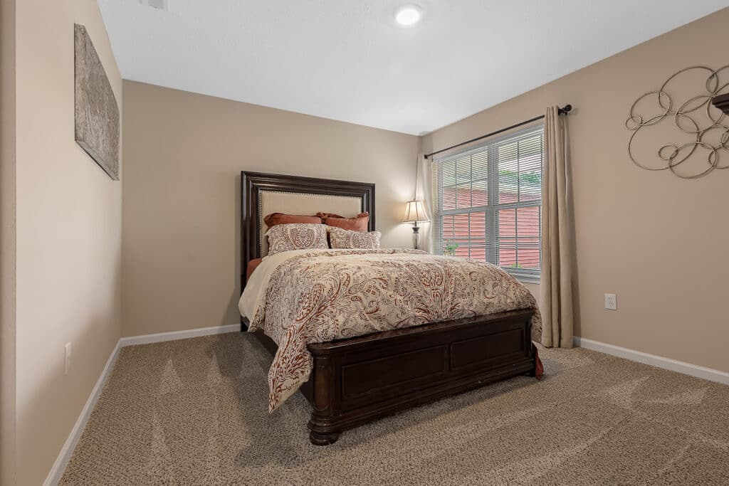 A neatly made bed with patterned bedding sits in a carpeted bedroom with beige walls, a large window with curtains, and wall decor.