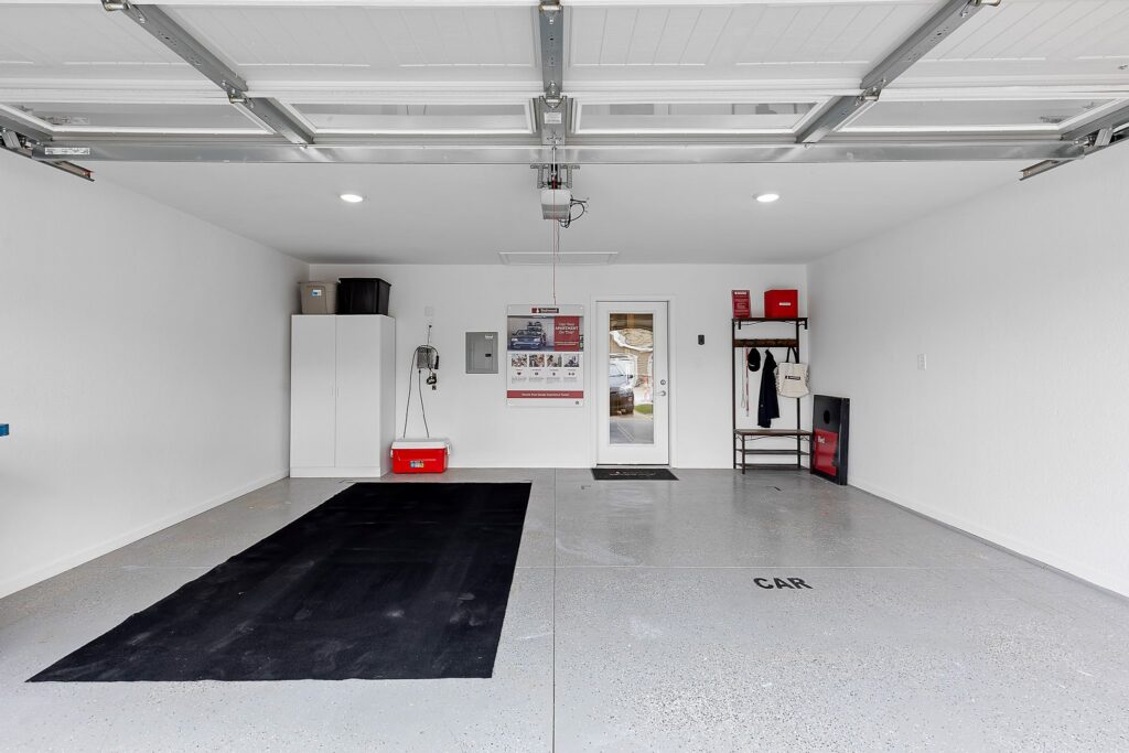 Clean, organized two-car garage with white walls, ceiling lights, storage cabinets, a wall-mounted coat rack, and a door leading outside. Black mat and floor markings label car spaces.
