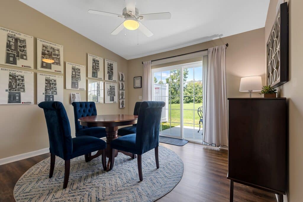 Dining area with a round table and four blue chairs on a rug, wall art, ceiling fan, cabinet, and sliding glass doors leading to a patio with outdoor view.