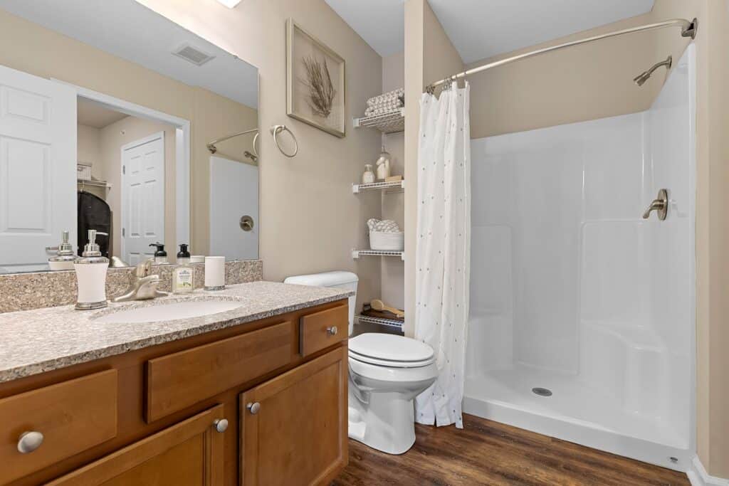 Bathroom with a granite countertop vanity, single sink, toilet, shelving with towels, and a walk-in shower with a white curtain. Neutral color scheme and wood-look flooring.