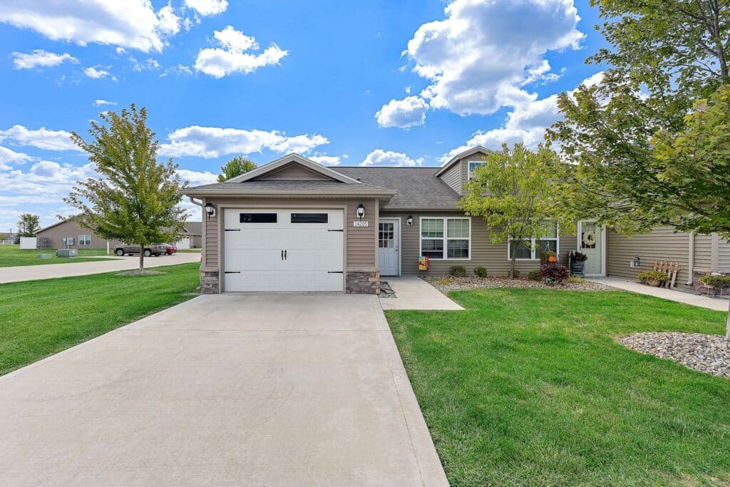 Single-story suburban house with attached garage, concrete driveway, green lawn, and a few trees under a partly cloudy sky.
