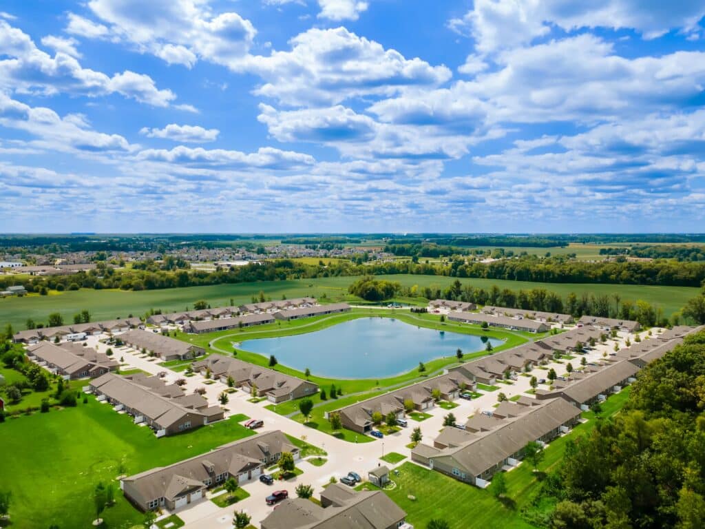 Aerial view of a suburban neighborhood with rows of houses surrounding a large pond, bordered by green fields and under a partly cloudy sky.