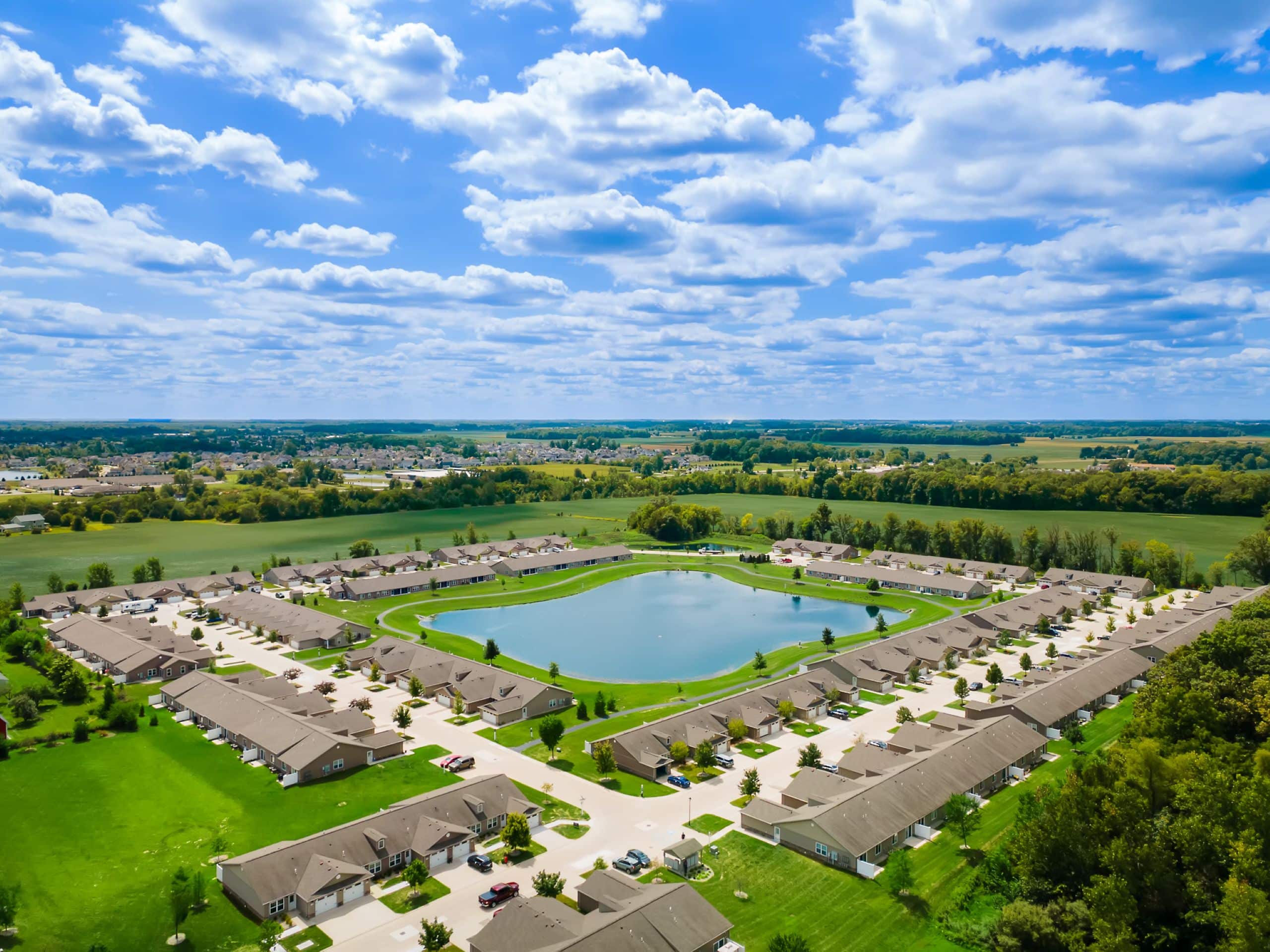 Aerial view of a suburban neighborhood with rows of houses surrounding a large pond, bordered by green fields and under a partly cloudy sky.