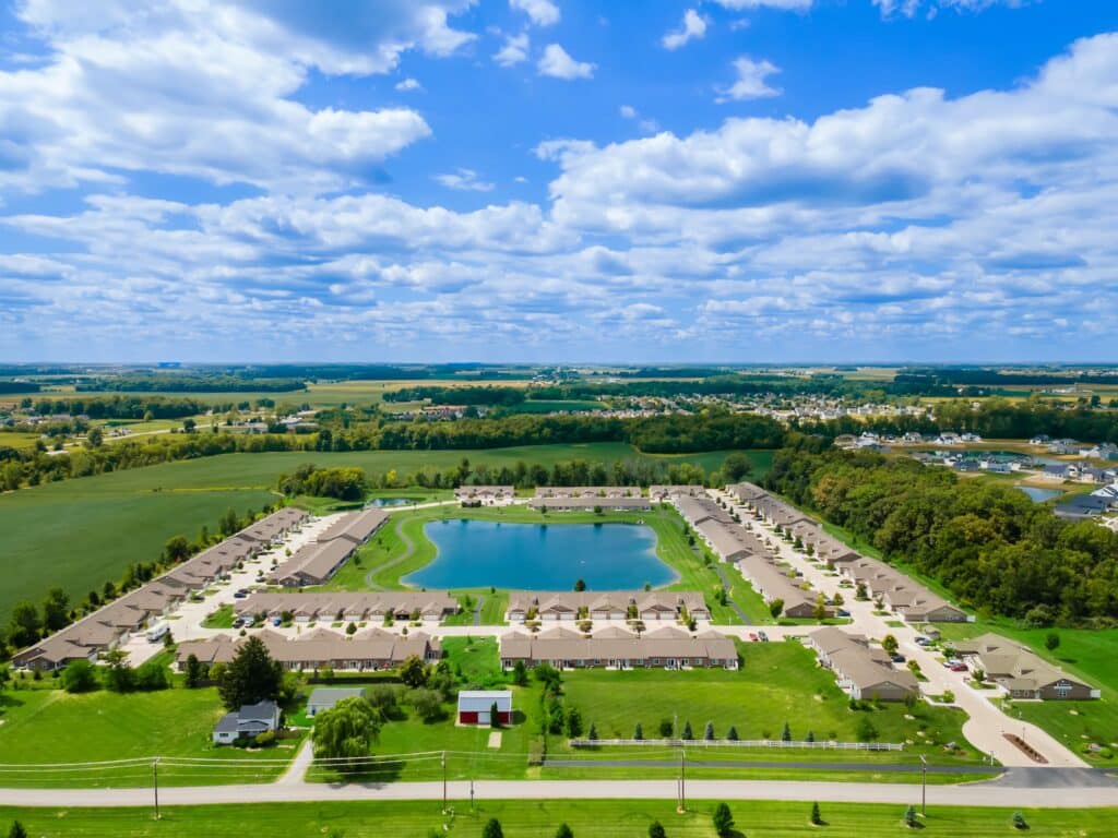 Aerial view of a residential neighborhood built around a central pond, surrounded by green fields and trees under a partly cloudy sky.