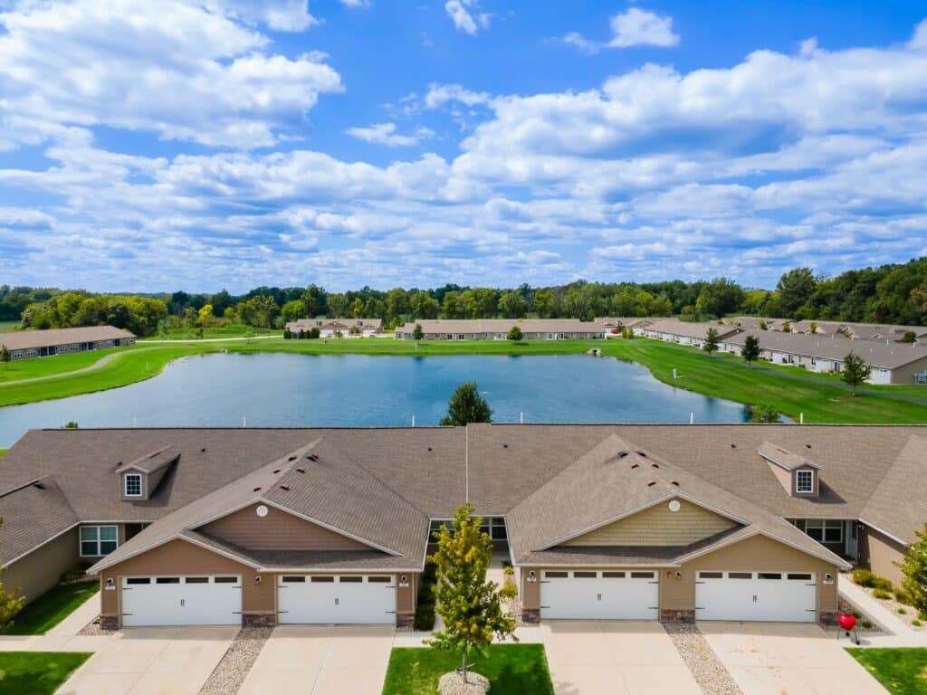 A row of tan townhouses with garages faces a large pond surrounded by green lawns, trees, and similar buildings under a partly cloudy sky.