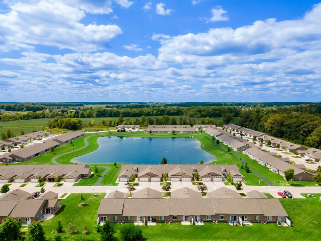 Aerial view of a suburban neighborhood with single-story houses surrounding a pond, green lawns, trees, and a partly cloudy sky.