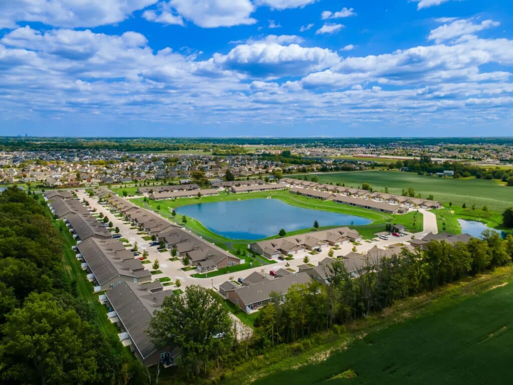 Aerial view of a suburban neighborhood with rows of houses surrounding a small artificial lake, bordered by green fields and trees under a blue sky with scattered clouds.