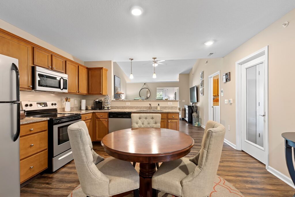 Contemporary kitchen and dining area with wooden cabinets, stainless steel appliances, round dining table, and upholstered chairs on hardwood flooring.