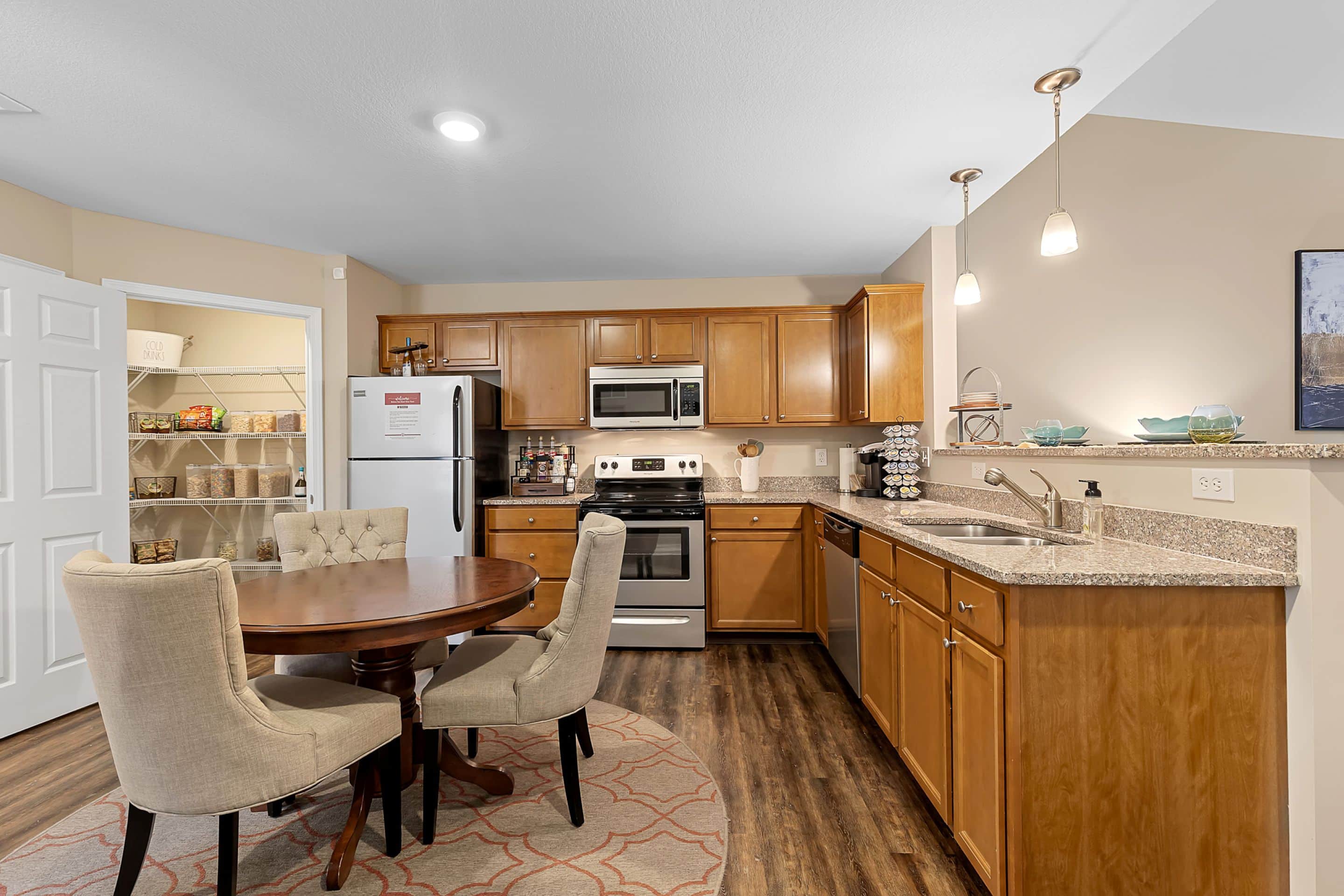 Modern kitchen with wooden cabinets, stainless steel appliances, round dining table with two chairs, pantry with shelves, and pendant lights above the sink.