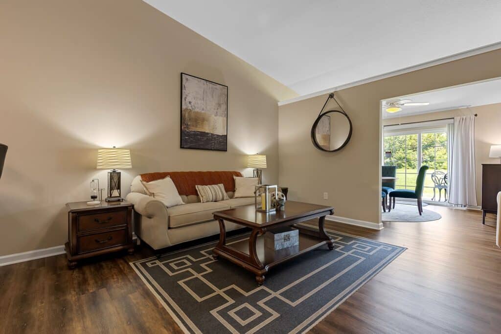 A living room with a beige sofa, two side tables with lamps, a wooden coffee table, patterned rug, wall art, and a round mirror, opening to a dining area with large windows.