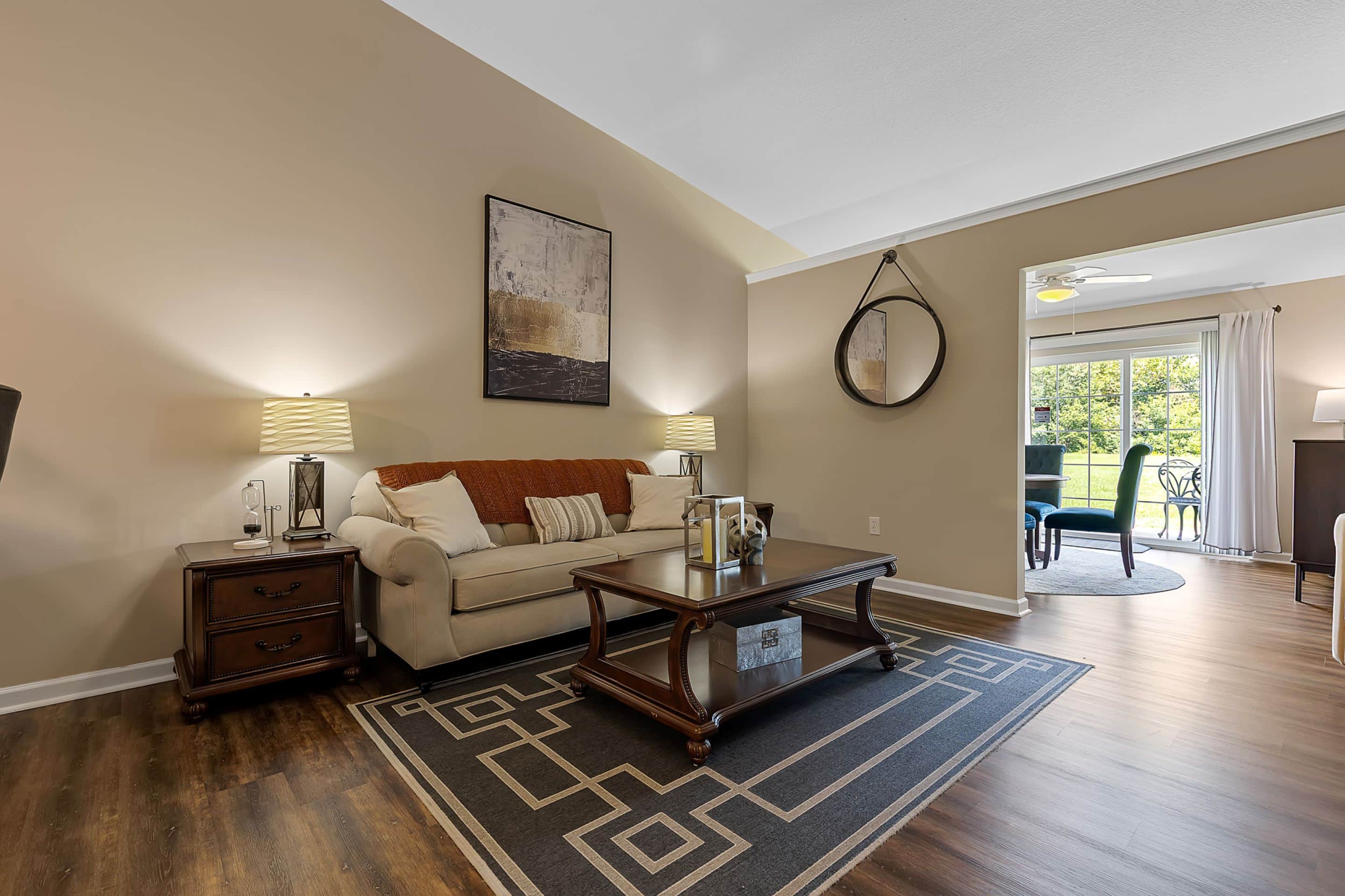 A living room with a beige sofa, two side tables with lamps, a wooden coffee table, patterned rug, wall art, and a round mirror, opening to a dining area with large windows.