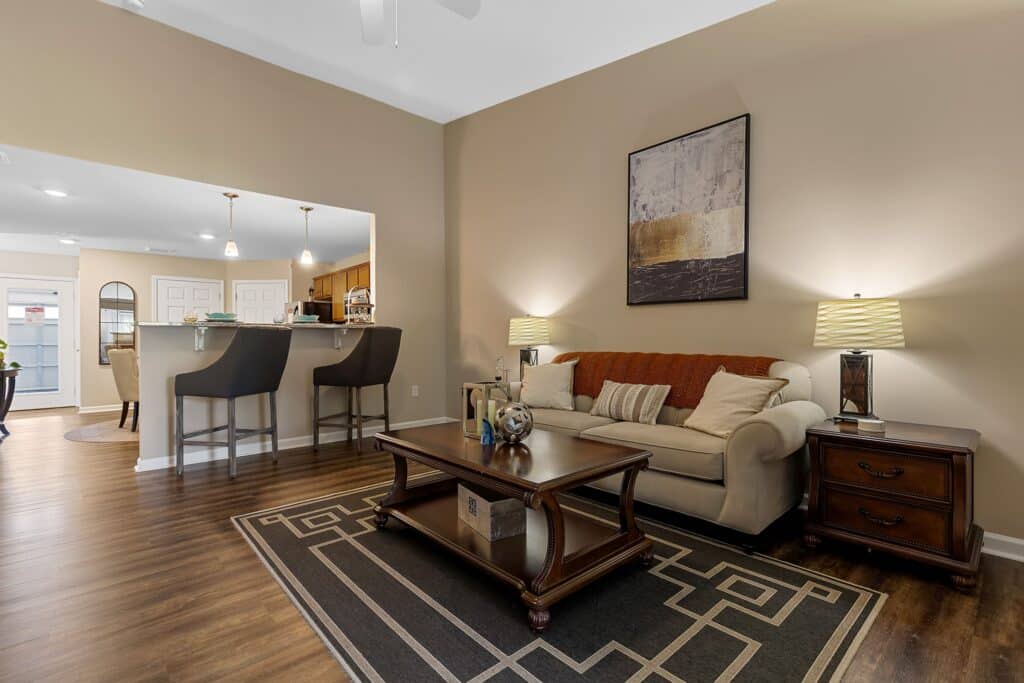A living room with a beige sofa, dark wood coffee table, two matching lamps, and a geometric rug, adjacent to a kitchen with bar stools and a breakfast bar.