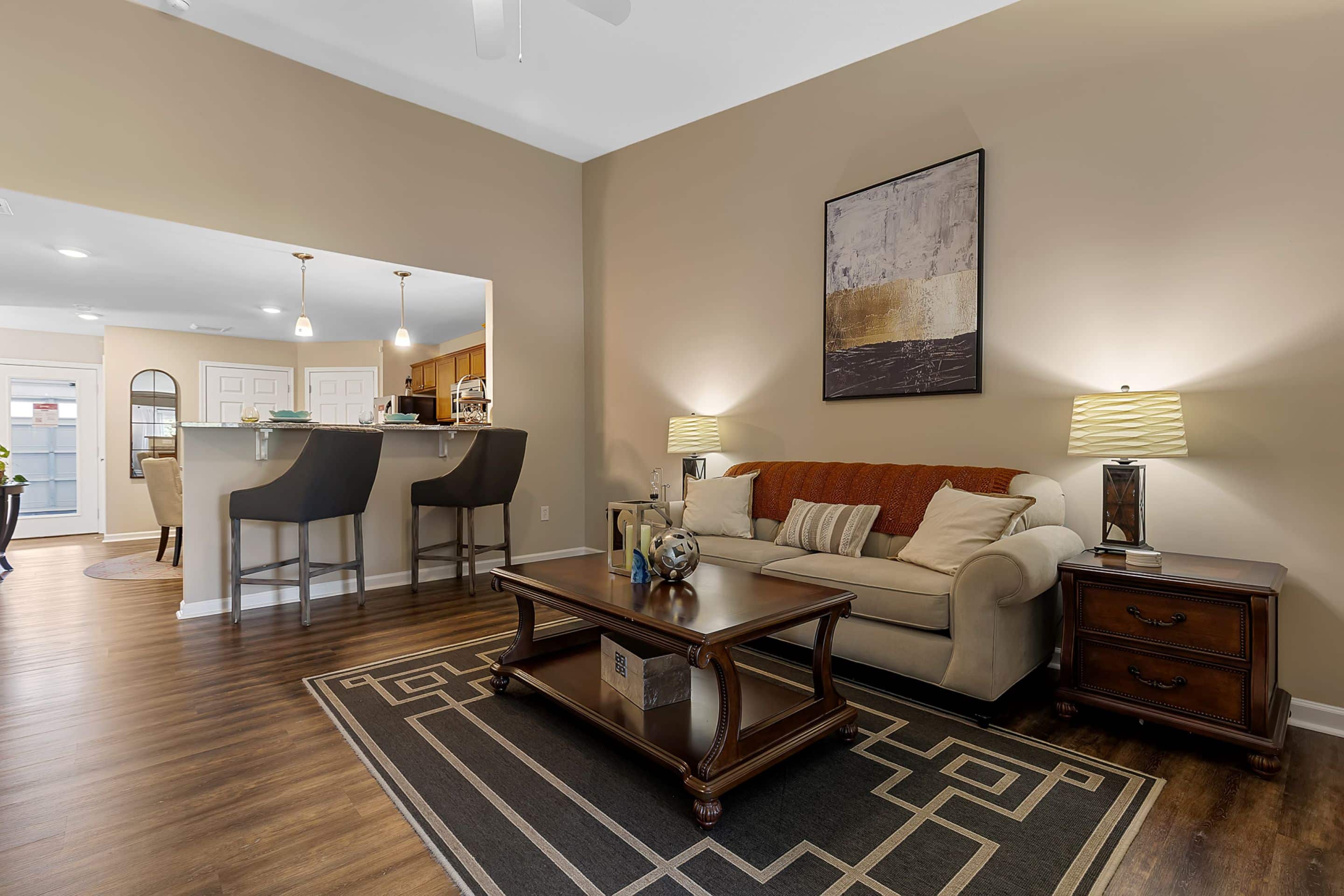 A living room with a beige sofa, dark wood coffee table, two matching lamps, and a geometric rug, adjacent to a kitchen with bar stools and a breakfast bar.