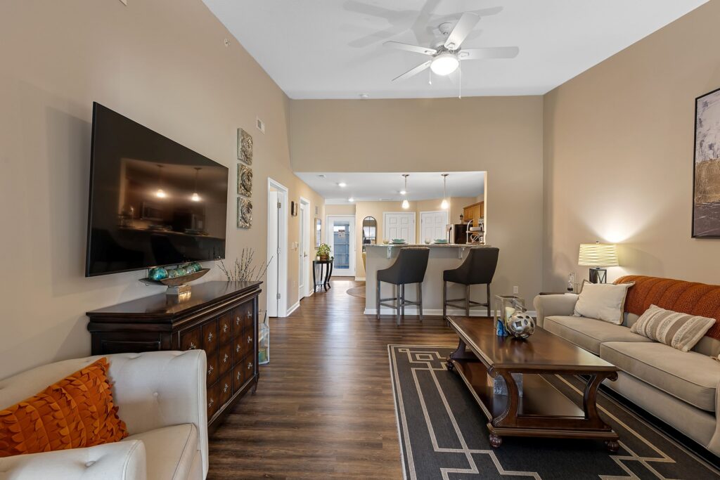 A modern living room with a mounted TV, beige furniture, a wooden coffee table, and an open view into the kitchen with bar stools and pendant lighting.