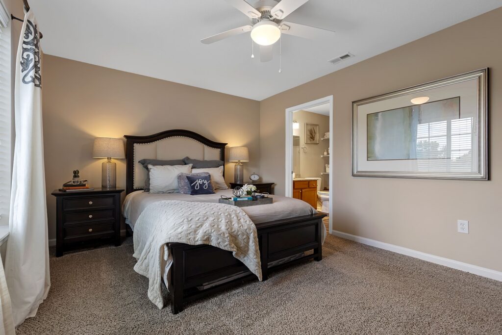 A neatly made bed with dark wood frame, nightstands with lamps, carpeted floor, wall art, and an open door leading to a bathroom in a beige-walled bedroom.