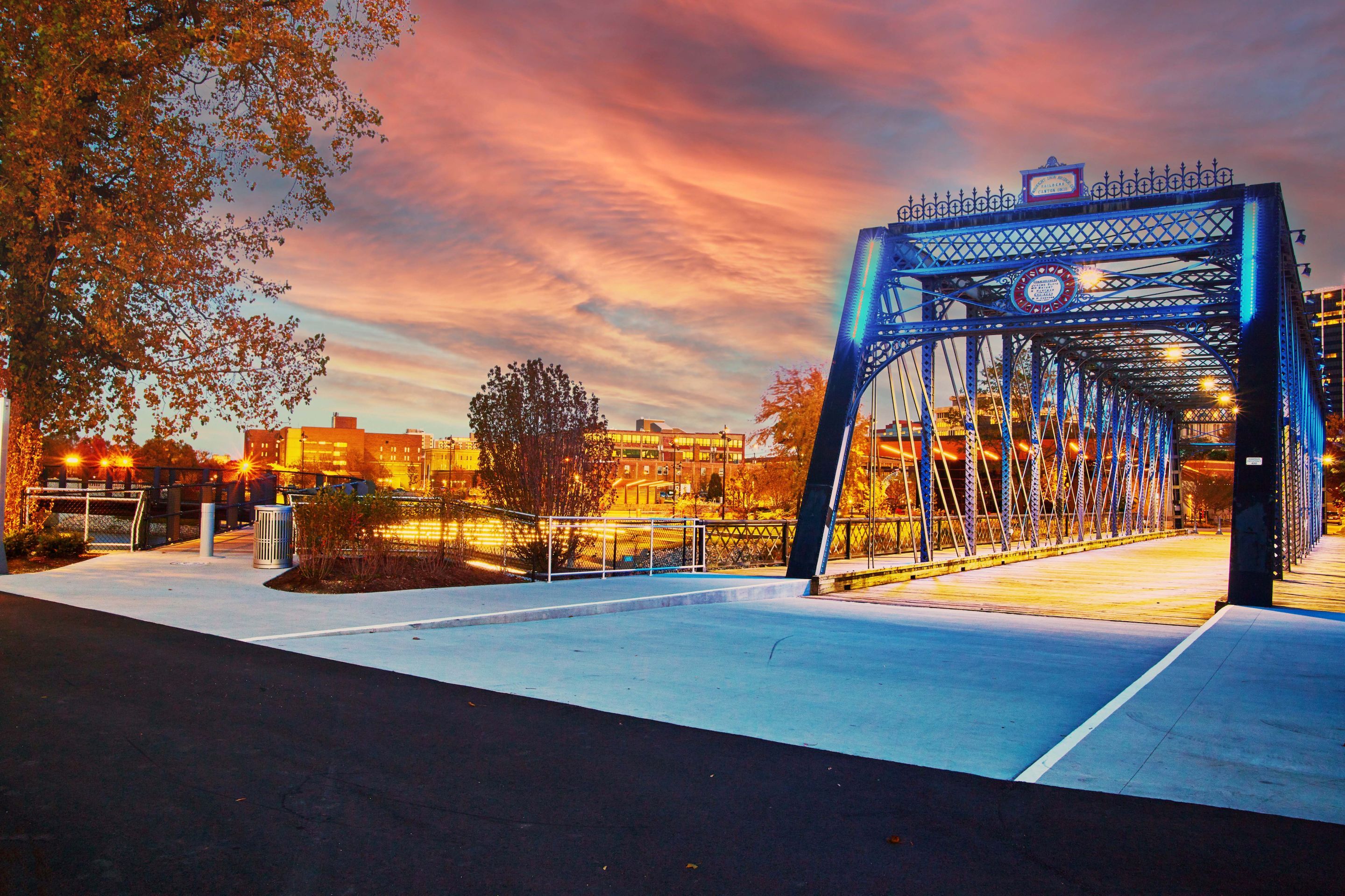 Blue steel truss bridge illuminated at dusk with city buildings and vibrant sunset sky in the background.