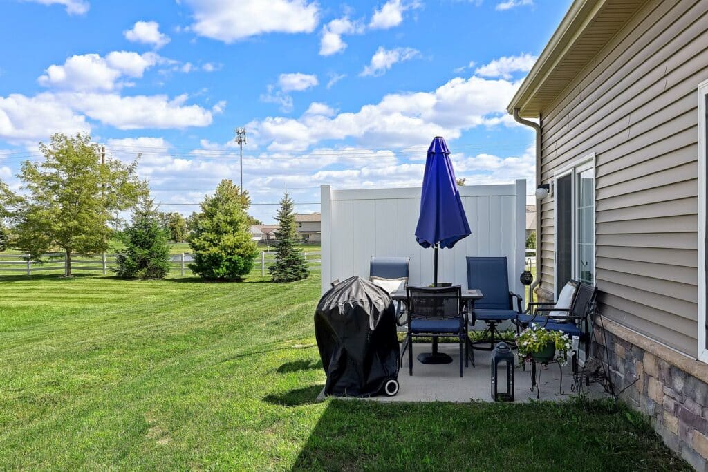 A backyard patio with a grill, table, chairs, blue umbrella, and potted plants beside a beige house; green lawn and trees in the background under a partly cloudy sky.