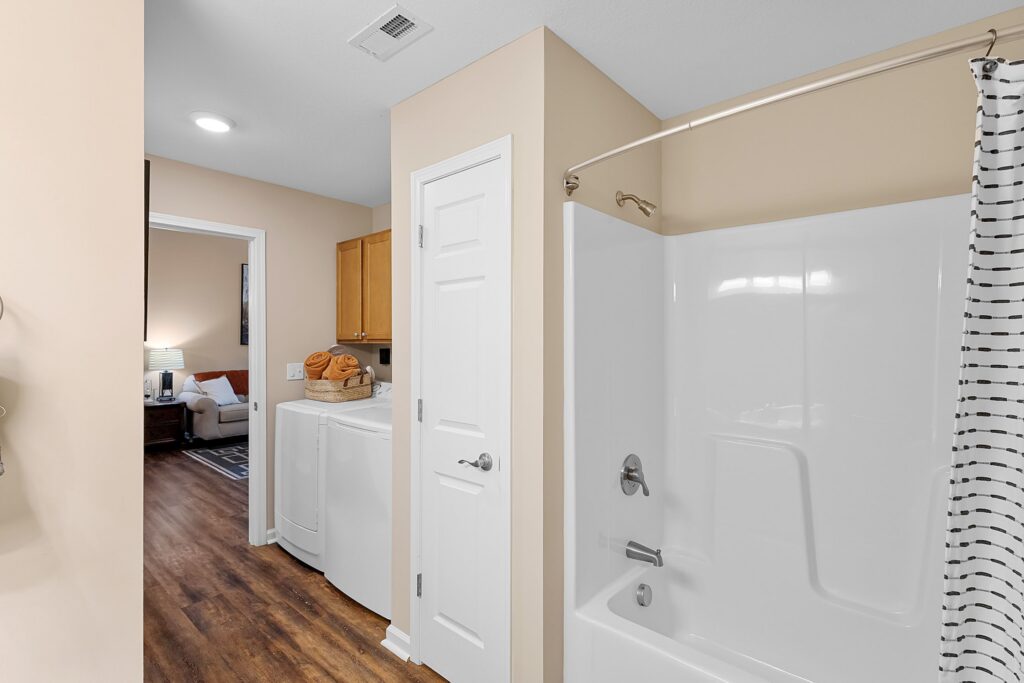 Bathroom with a white bathtub and shower curtain, adjoining a laundry area with a washer, dryer, and wooden cabinets, visible through an open doorway.