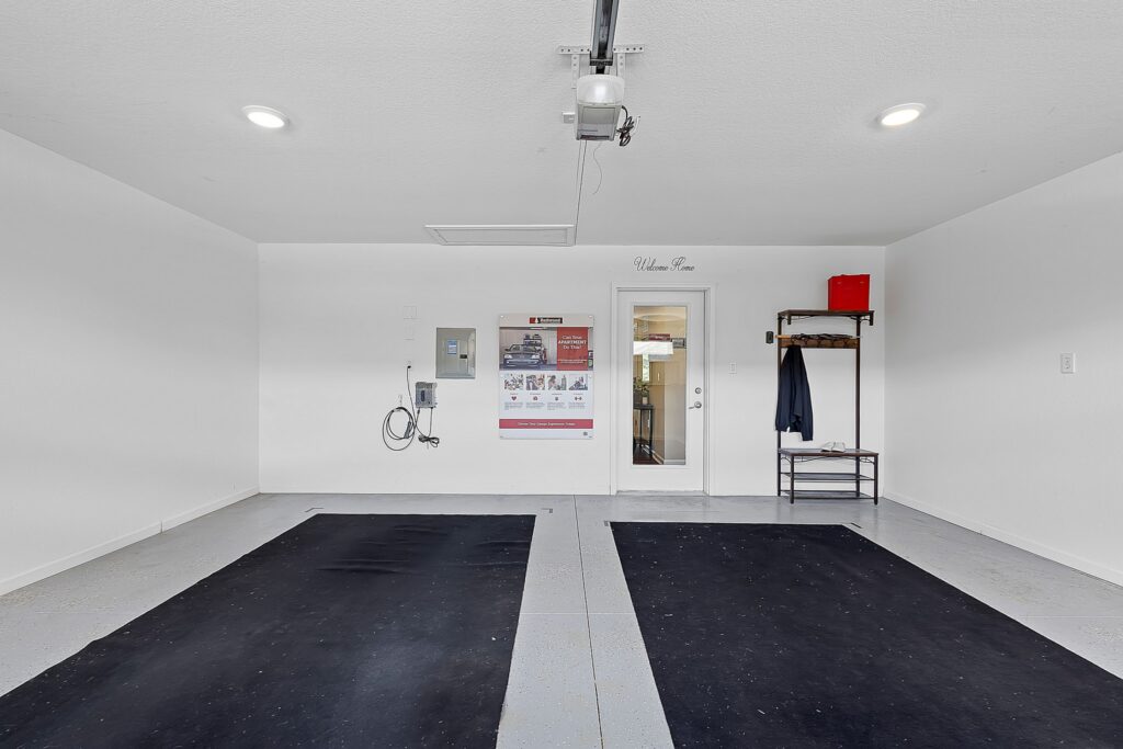 Clean, empty garage with two black mats on the floor, a wall-mounted device, a rack with hangers and a red box, and a door leading inside. White walls and ceiling, bright lighting.