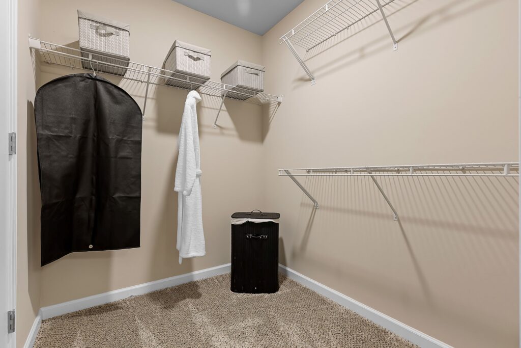 Empty walk-in closet with beige walls, wire shelving, a black garment bag, a white robe, storage baskets, and a black laundry hamper on a carpeted floor.