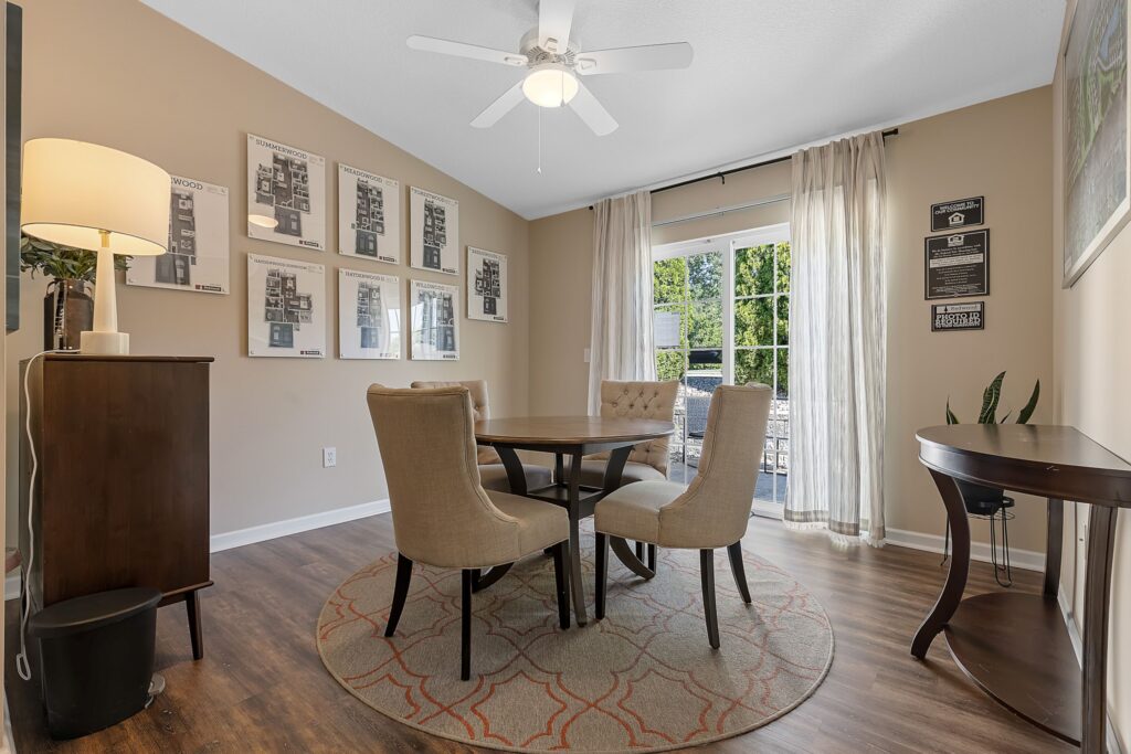 A dining room with a round table and four chairs on a rug, wood flooring, framed art on the walls, a side table, a cabinet, and a sliding glass door with curtains.
