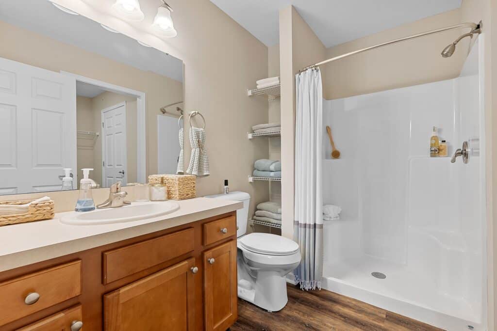 Bathroom with wooden vanity, single sink, toilet, open shelving with towels, walk-in shower with curtain, and light fixtures above the mirror.
