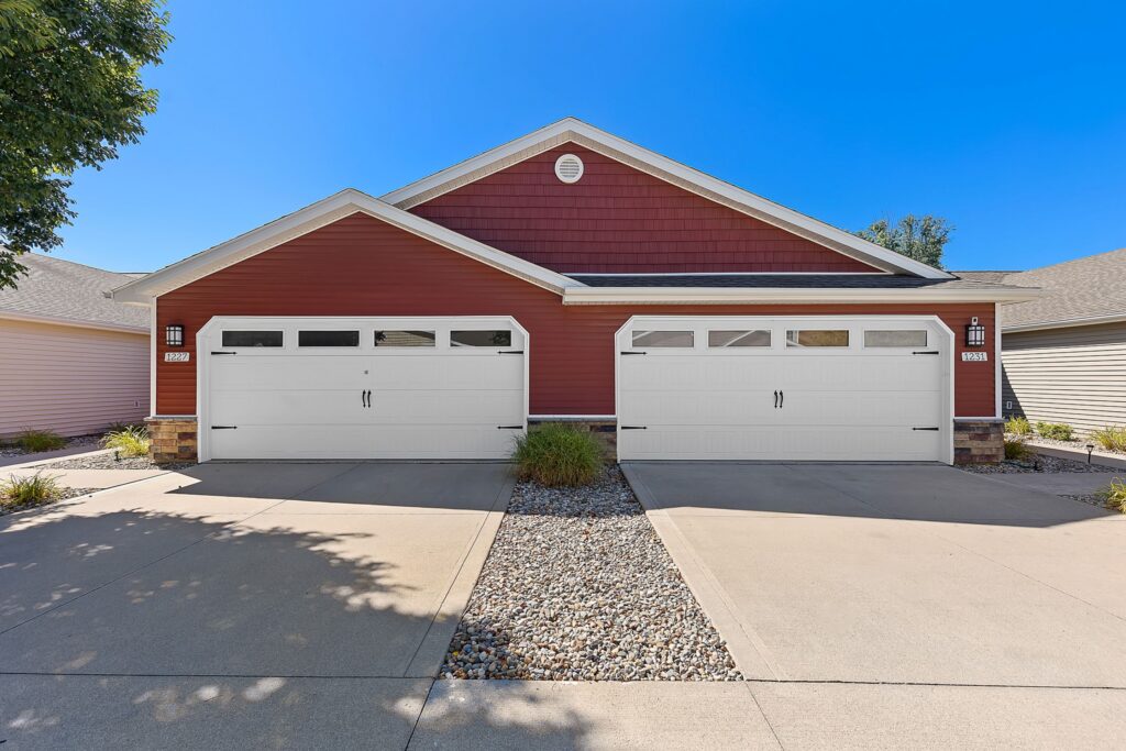 A duplex with red siding and white trim, each unit featuring a two-car garage, concrete driveways, and a stone-lined center divider with small plants.
