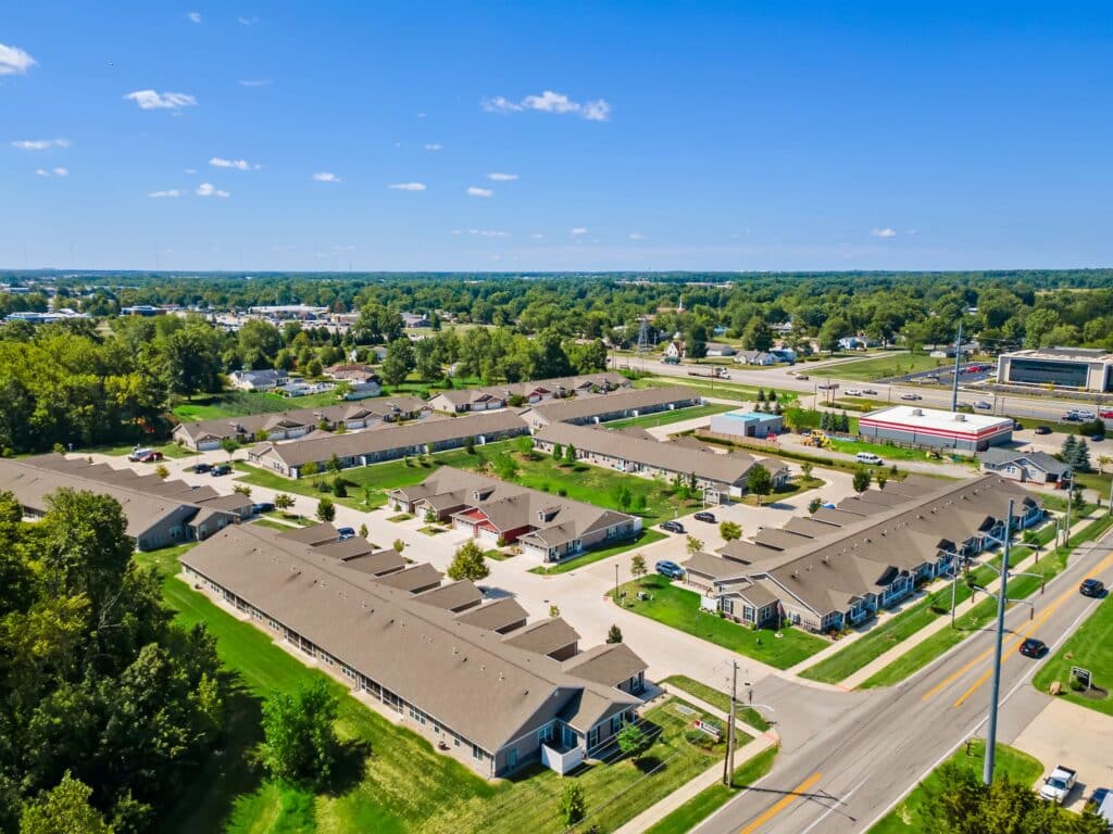 Aerial view of a residential neighborhood with single-story houses, paved roads, driveways, green lawns, and surrounding trees under a clear blue sky.