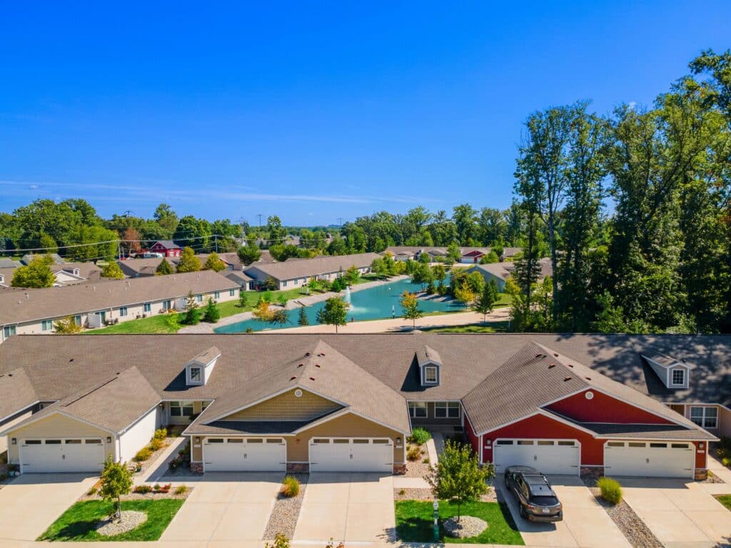 A row of attached suburban houses with garages, a parked car, and landscaped lawns sits in front of a neighborhood pond surrounded by more homes and trees under a clear blue sky.
