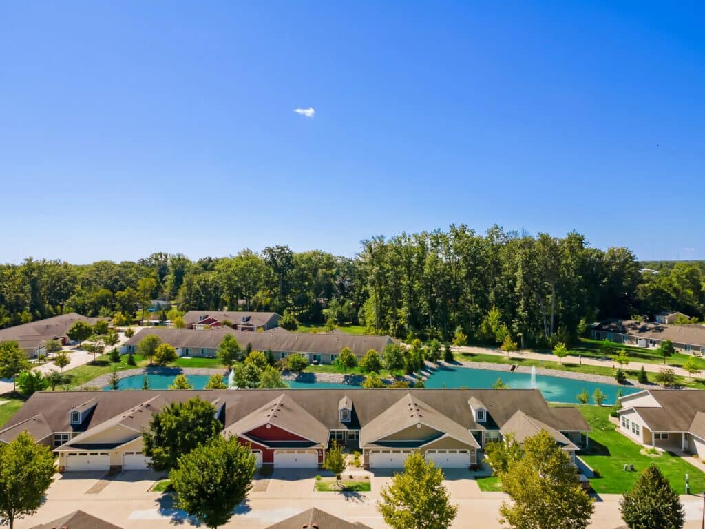 Aerial view of a suburban neighborhood with rows of houses, green lawns, trees, and a small pond under a clear blue sky.