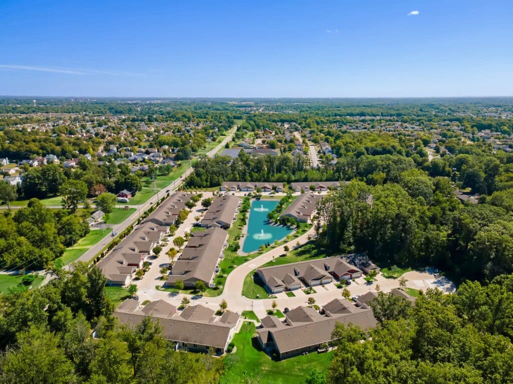Aerial view of a suburban neighborhood with rows of houses surrounding a central pond, trees, and green spaces under a clear blue sky.