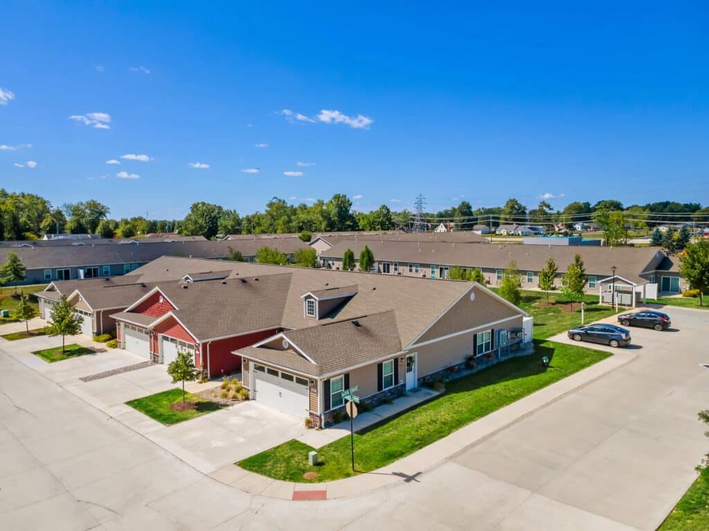 Aerial view of a suburban neighborhood with single-story houses, driveways, parked cars, green lawns, and clear blue sky.