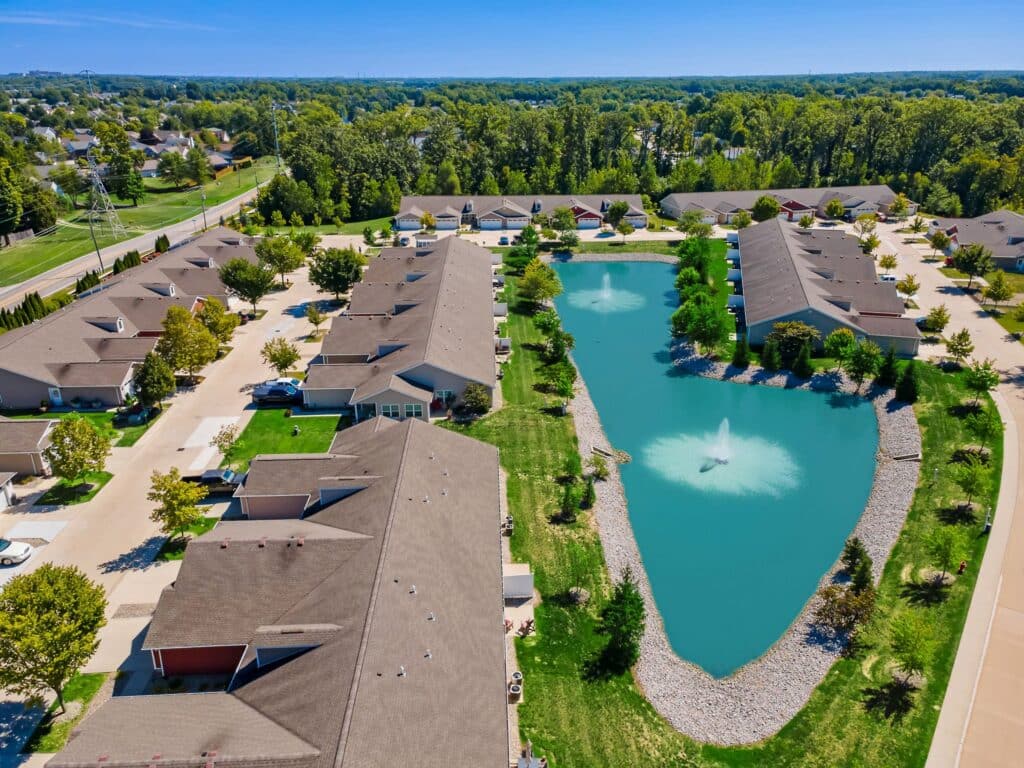 Aerial view of a residential neighborhood with rows of houses surrounding a pond with a central fountain, green lawns, and trees on a clear sunny day.